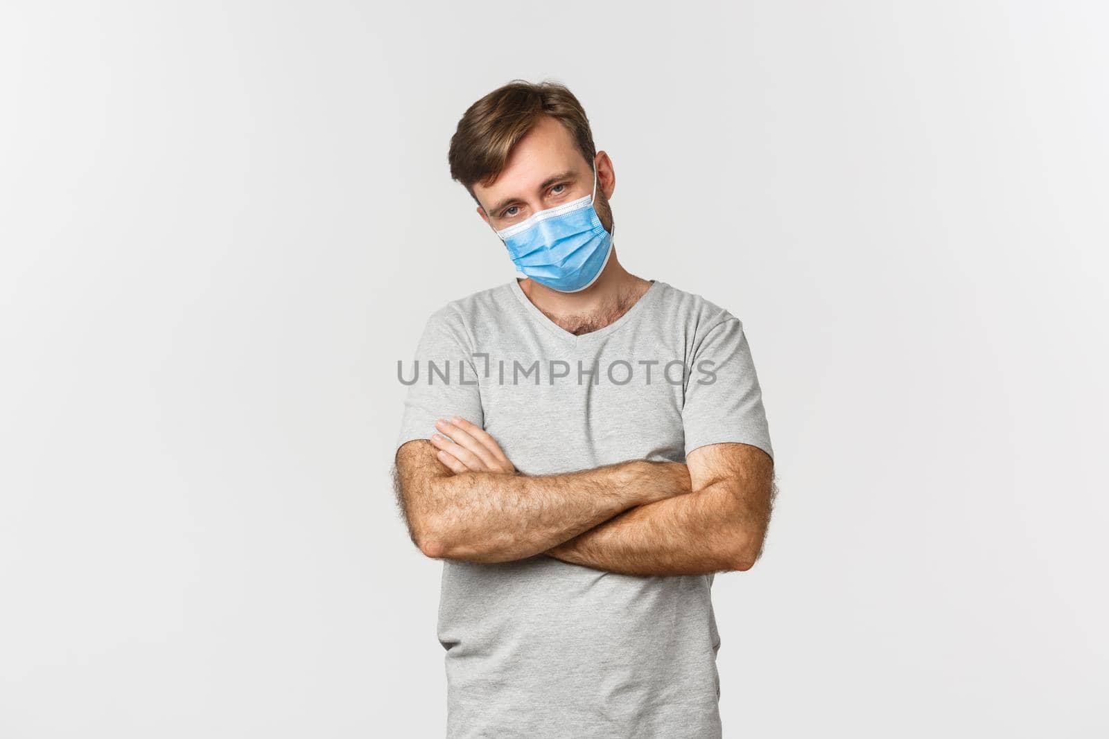 Concept of pandemic, covid-19 and social-distancing. Annoyed young man in gray t-shirt and medical mask, cross arms on chest and looking unamused, standing over white background.