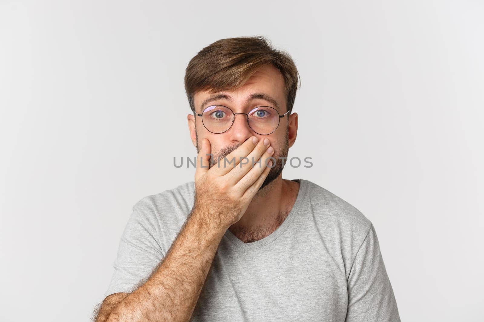 Close-up of shocked and worried man in glasses, gasping and cover mouth, hear something scary, standing over white background by Benzoix