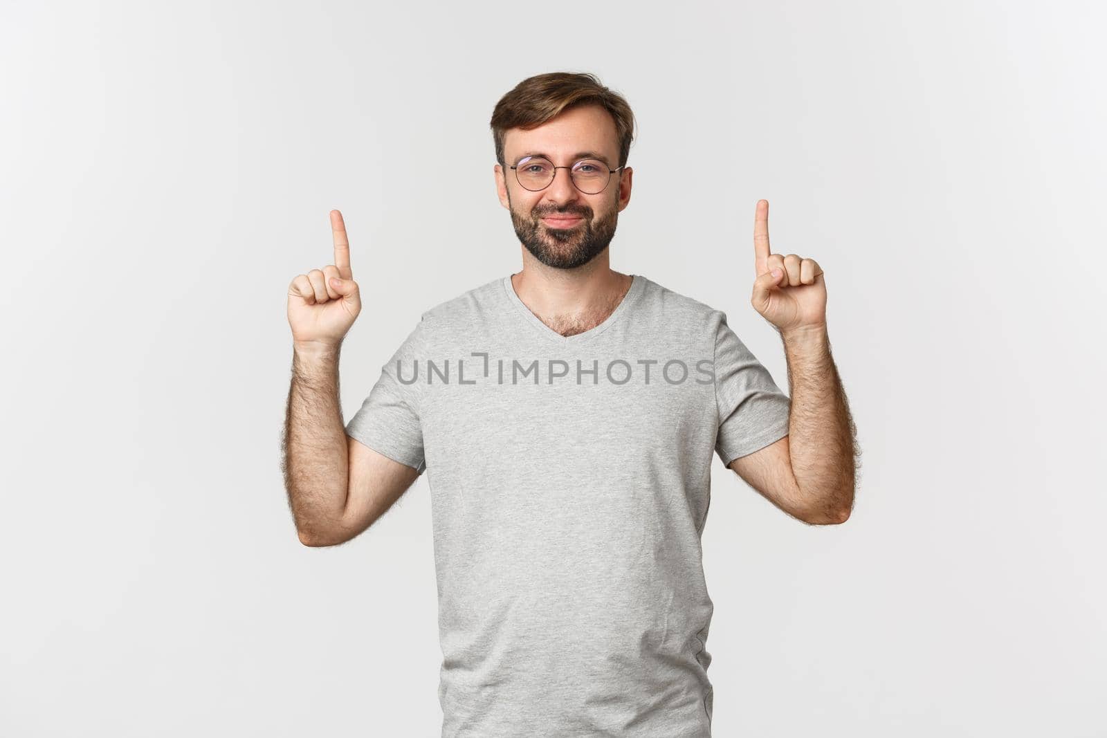 Cheerful bearded man smiling, pointing fingers up, showing logo, wearing gray t-shirt, standing over white background.