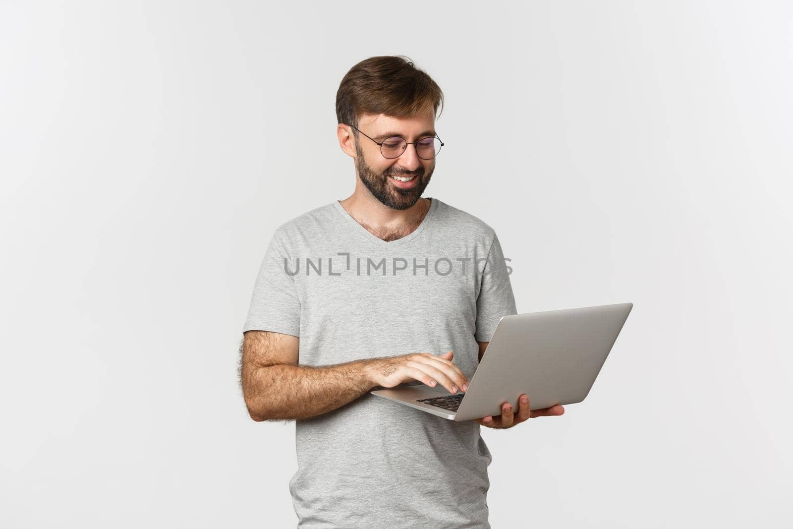 Handsome man freelancer, working with laptop and smiling, wearing glasses and t-shirt, standing over white background.