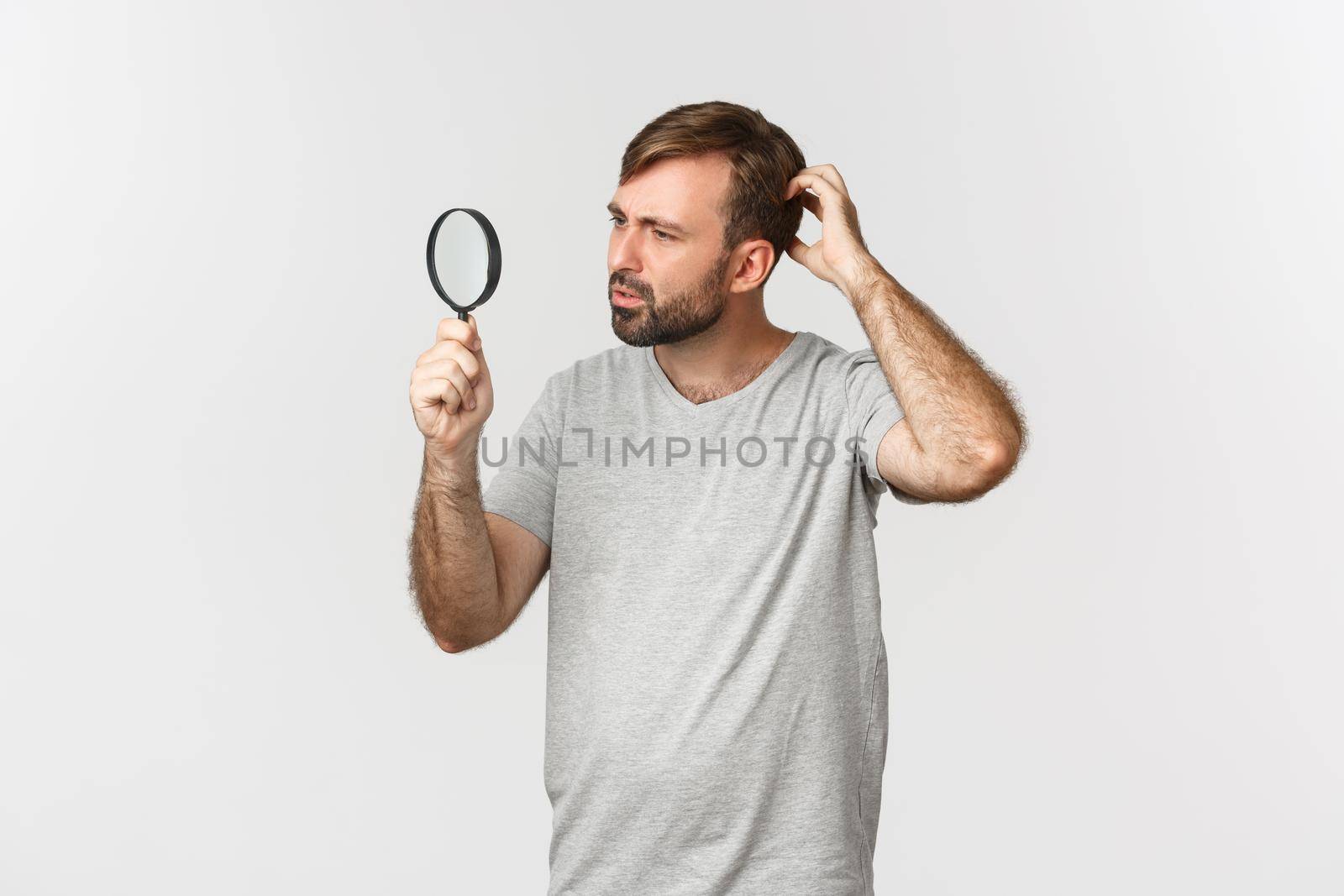 Image of handsome caucasian man in gray t-shirt, searching for something, looking confused through magnifying glass with curious face, standing over white background.