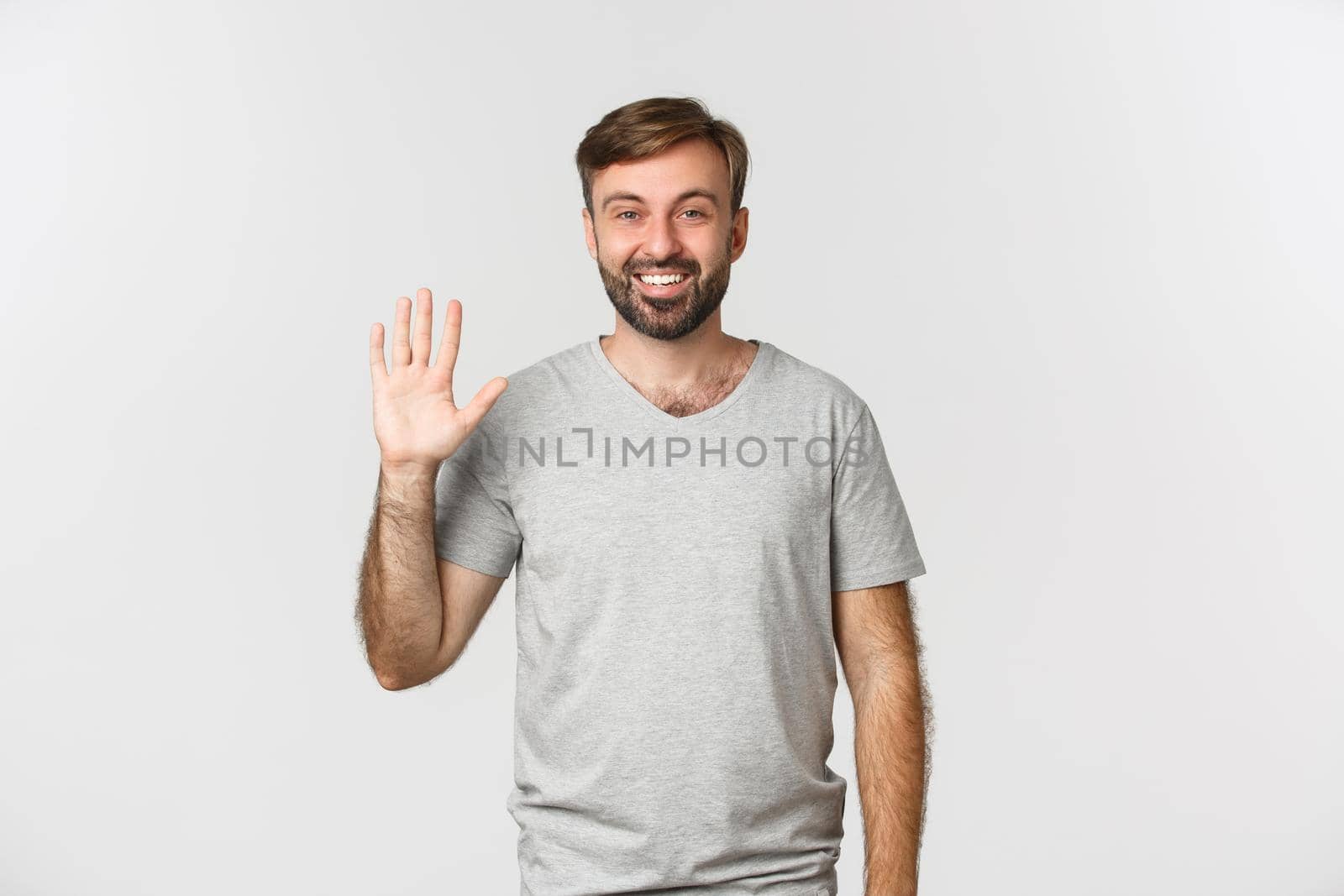 Portrait of handsome caucasian man in gray t-shirt, saying hello and friendly waving hand, standing over white background.