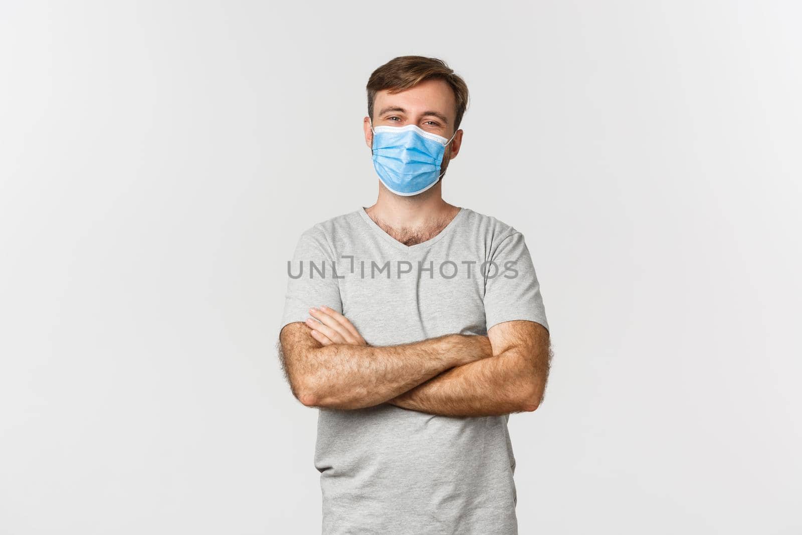 Concept of pandemic, covid-19 and social-distancing. Handsome and confident man in medical mask, cross arms on chest and smiling, standing over white background.