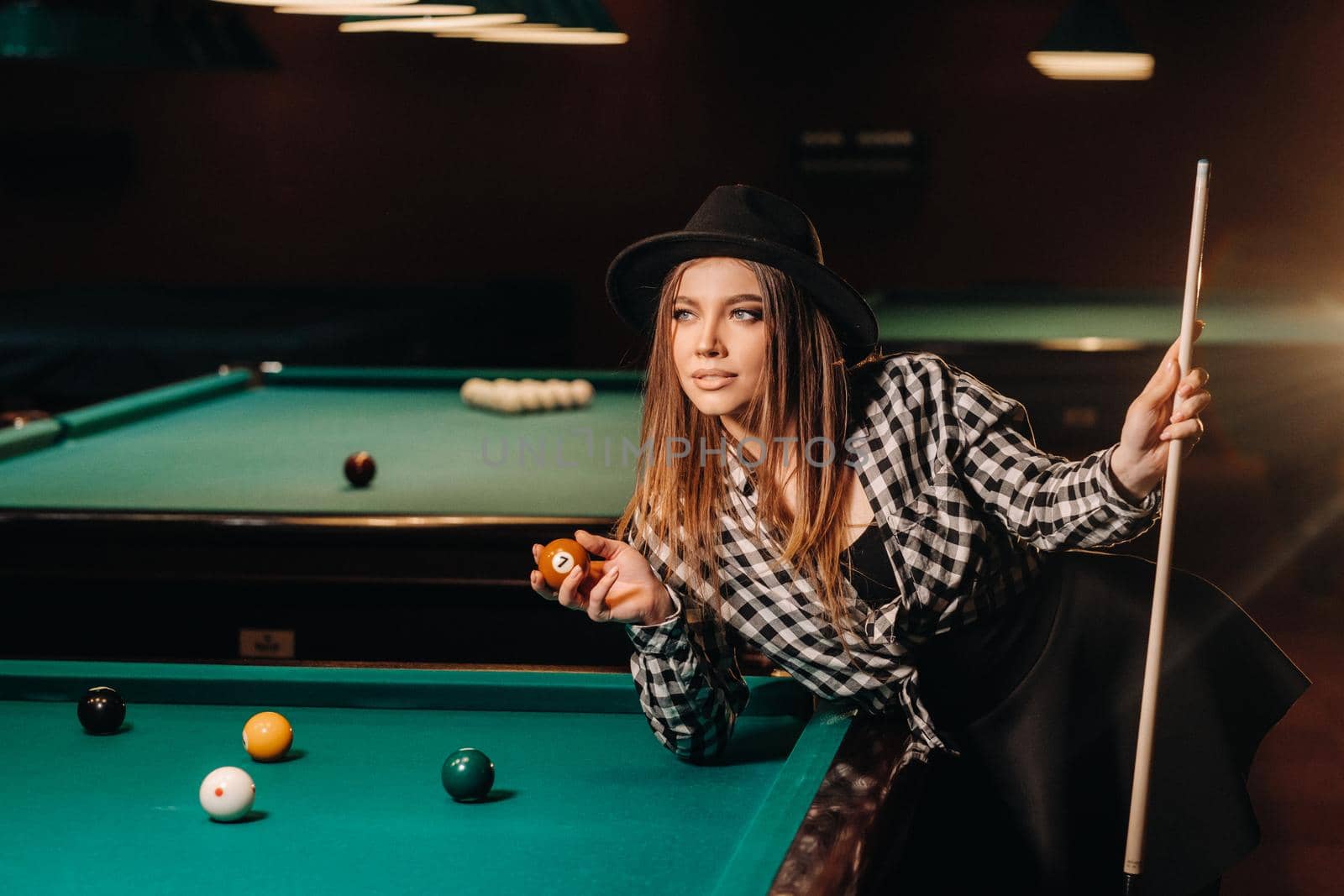 A girl in a hat in a billiard club with a cue and balls in her hands.Playing pool.