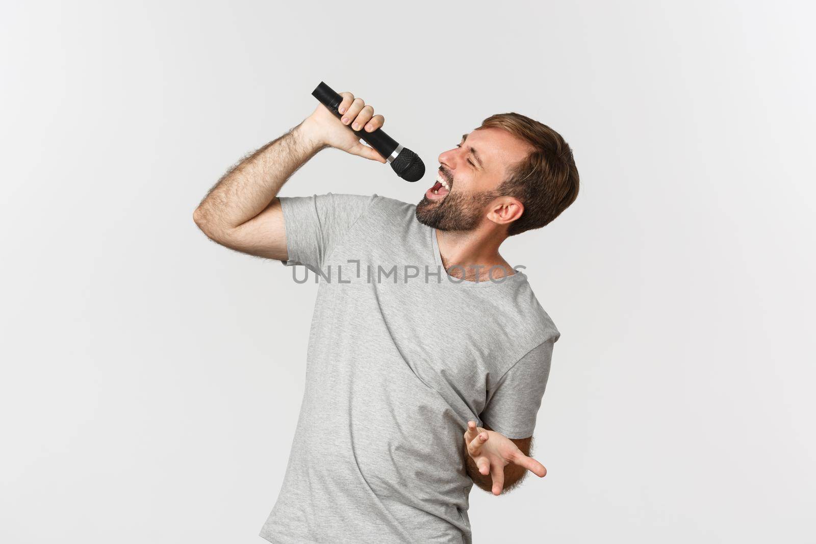 Handsome carefree guy singing song in karaoke, holding microphone, standing over white background.