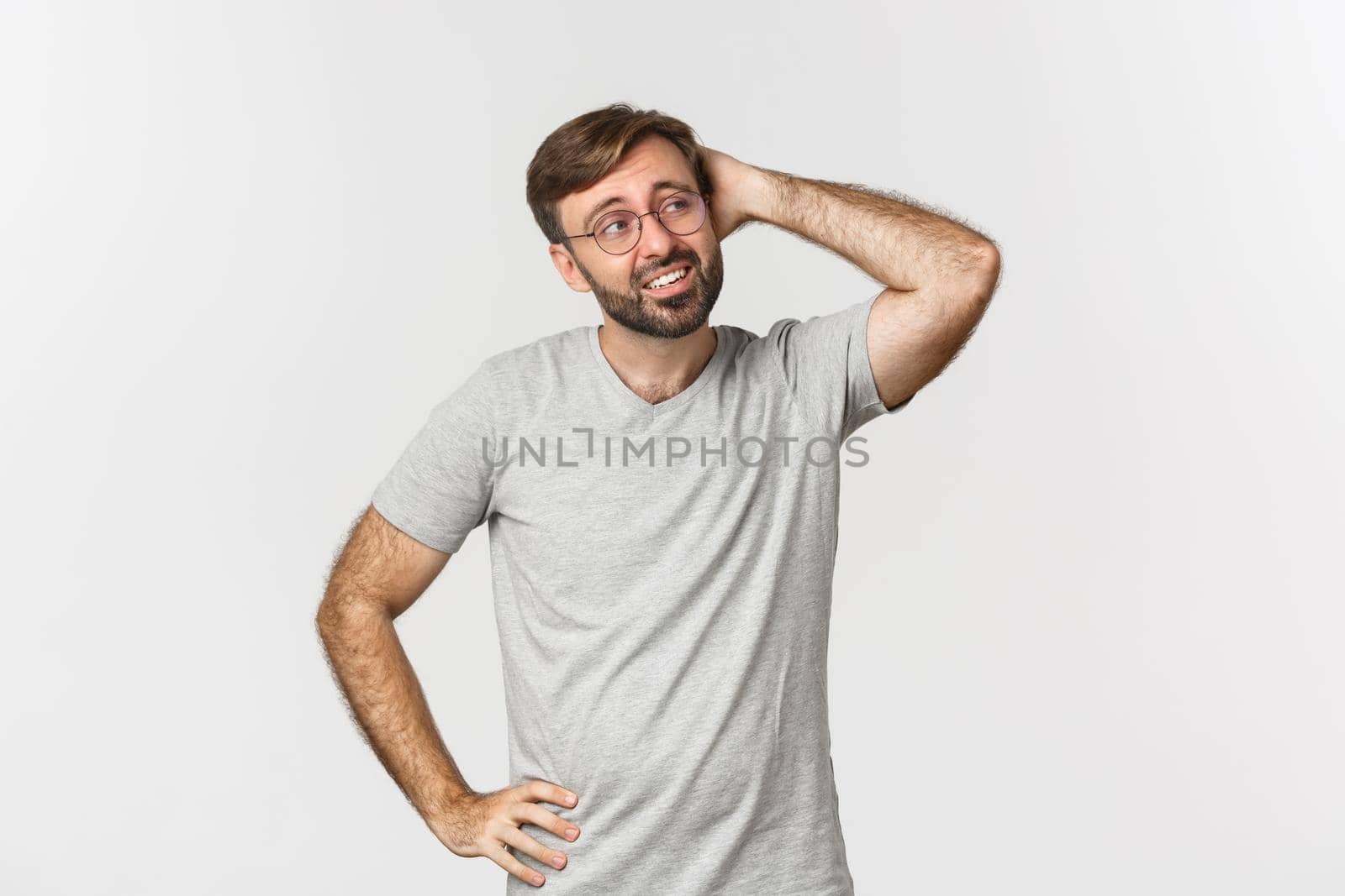 Portrait of indecisive adult man thinking, scratching head and looking confused at upper right corner, standing in gray t-shirt and glasses over white background.