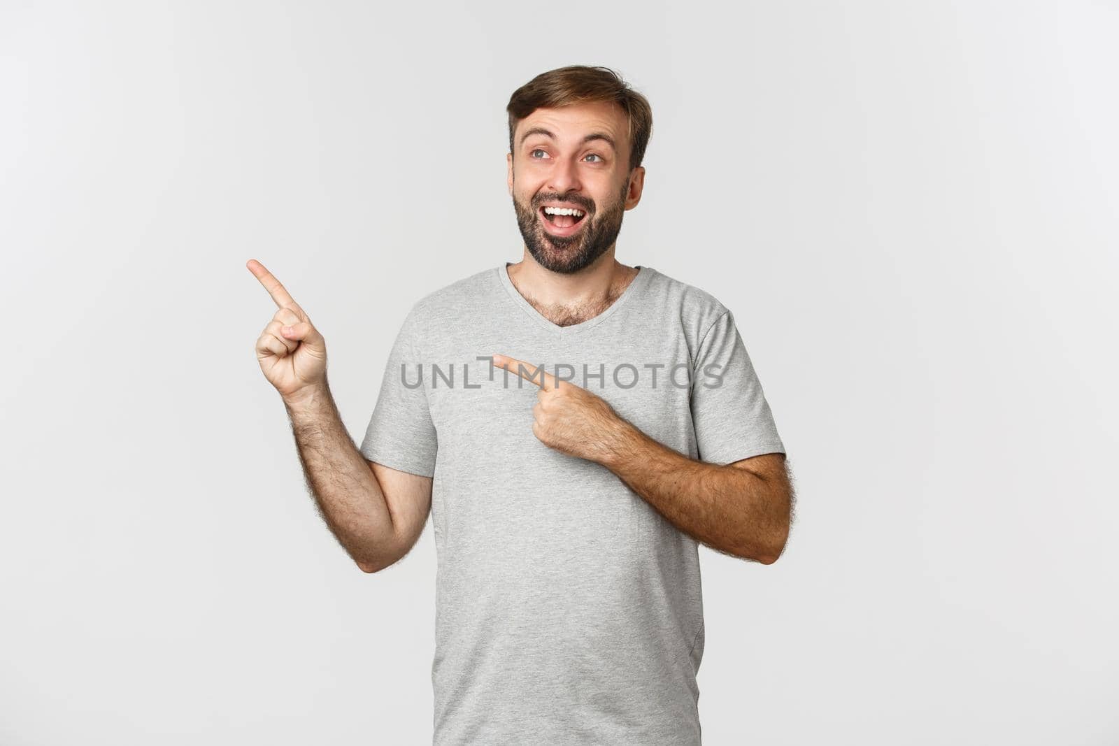 Handsome smiling man in gray t-shirt, looking happy and excited at upper left corner, showing logo, standing over white background.