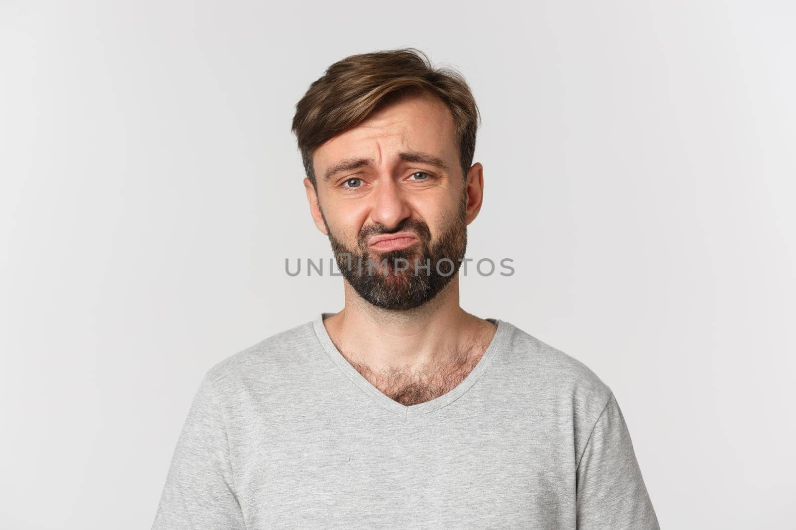 Close-up of gloomy disappointed guy with beard, wearing gray t-shirt, grimacing and frowning sad, standing over white background by Benzoix