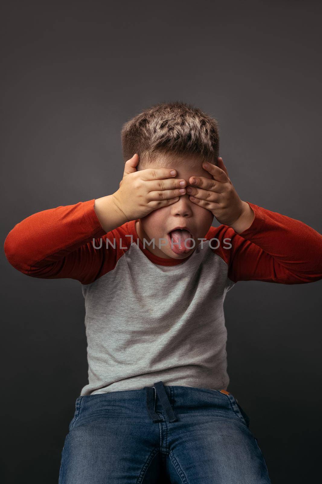 Little boy shows tongue with his eyes closed with hands. Preschooler expresses emotions in studio by LipikStockMedia