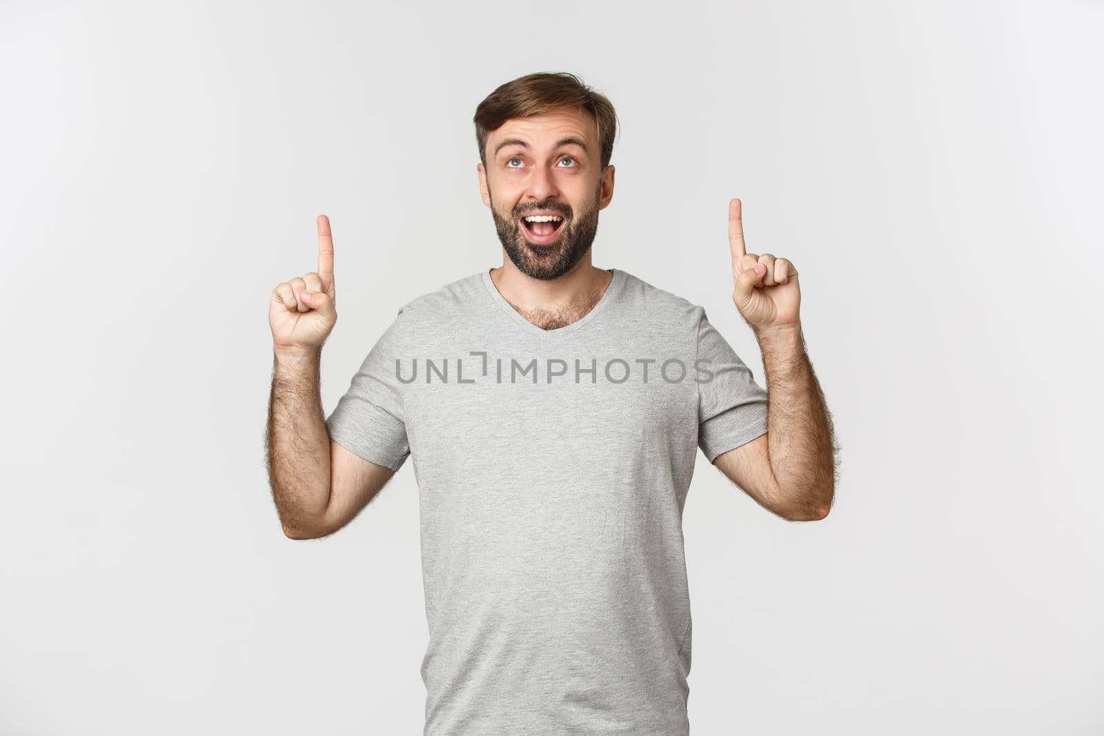 Portrait of handsome smiling man in gray t-shirt, looking and pointing fingers up with happy expression, showing logo, standing over white background by Benzoix