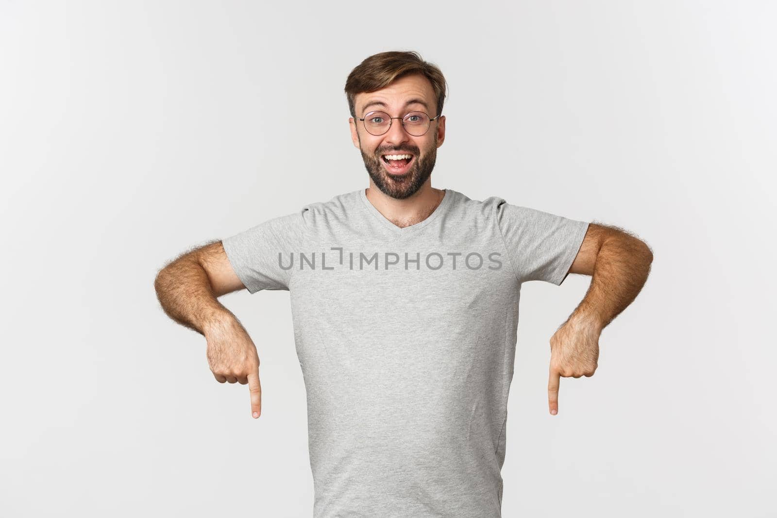 Cheerful bearded man smiling, pointing fingers down, showing logo, wearing gray t-shirt, wearing gray t-shirt, standing over white background.