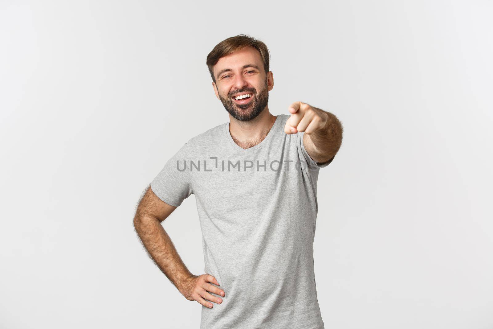 Image of carefree caucasian guy with beard, wearing gray t-shirt, laughing and pointing finger at camera, standing over white background.