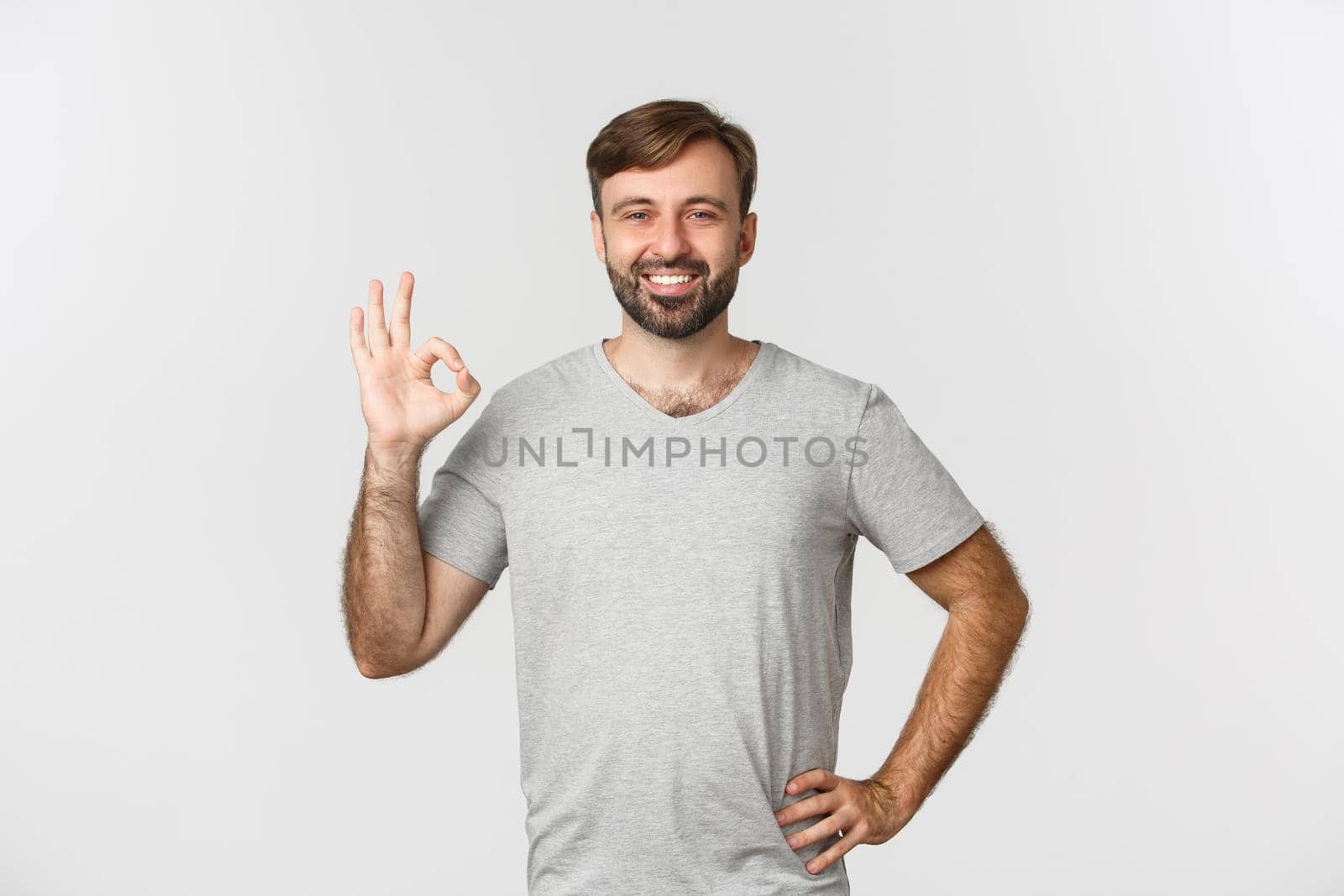 Portrait of handsome smiling man in casual grey t-shirt, showing okay sign in approval, agree to you, praising good choice, standing over white background.