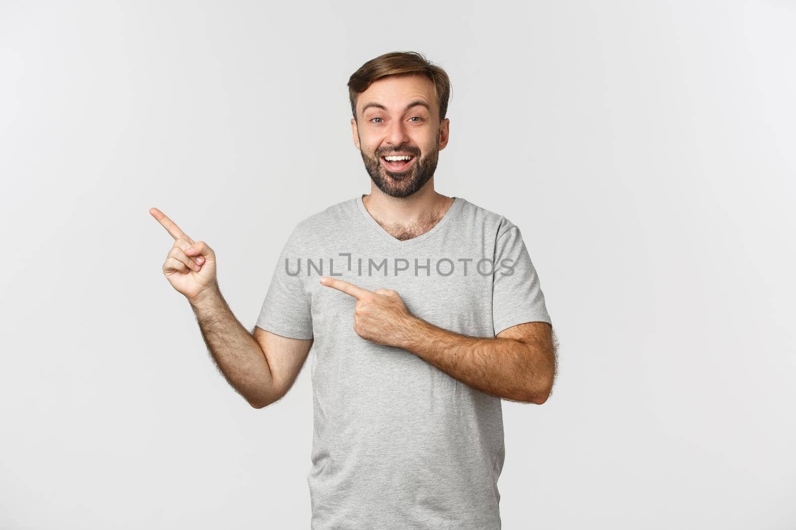 Portrait of excited smiling man with beard, wearing casual gray t-shirt, pointing fingers at upper left corner, showing logo, standing over white background.