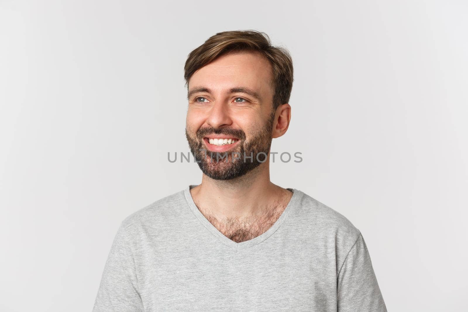 Portrait of handsome bearded man in gray t-shirt, looking left and smiling, standing over white background.