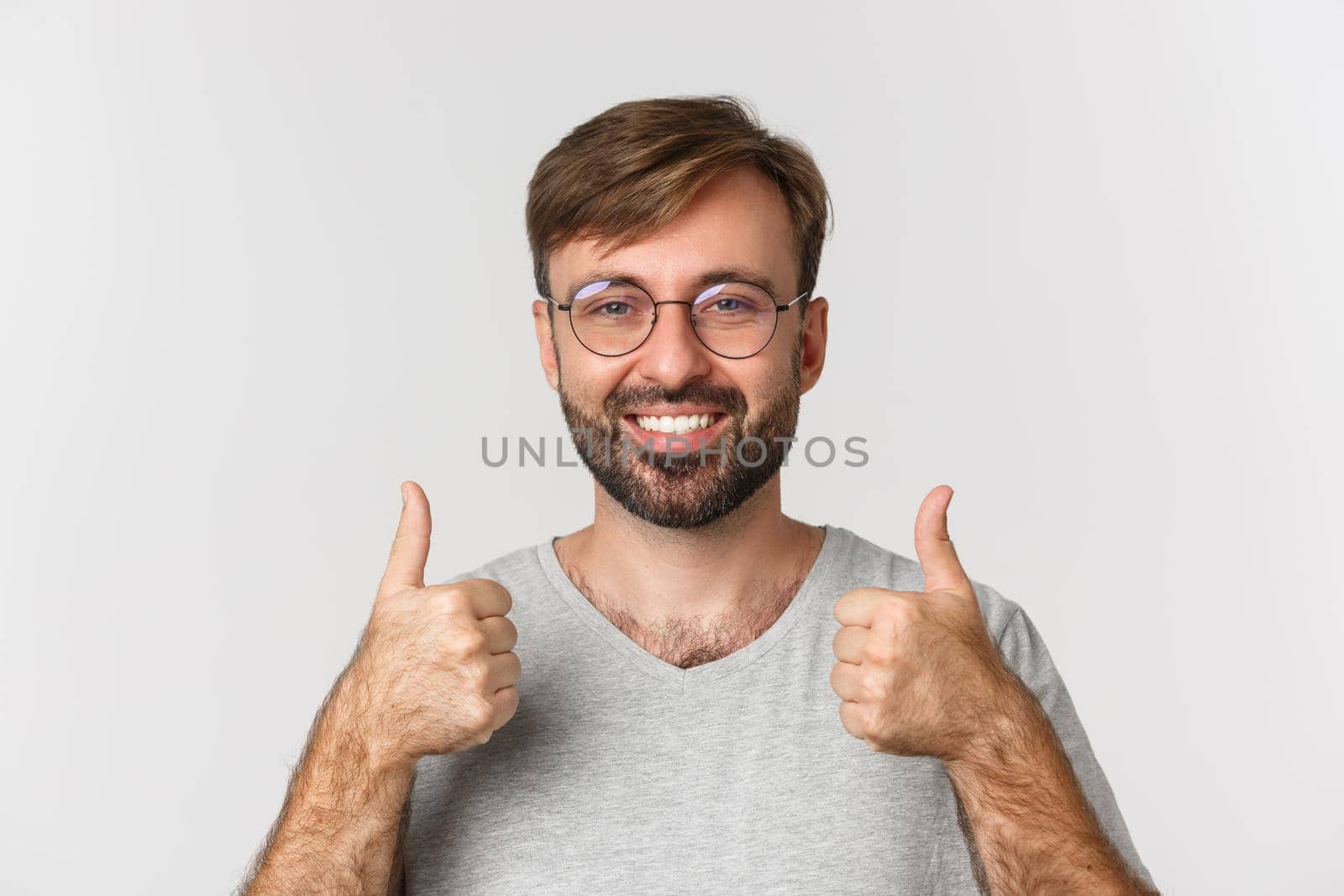 Close-up of cheerful caucasian man with beard, wearing glasses and casual t-shirt, smiling and showing thumbs-up in approval, recommend product, standing over white background.