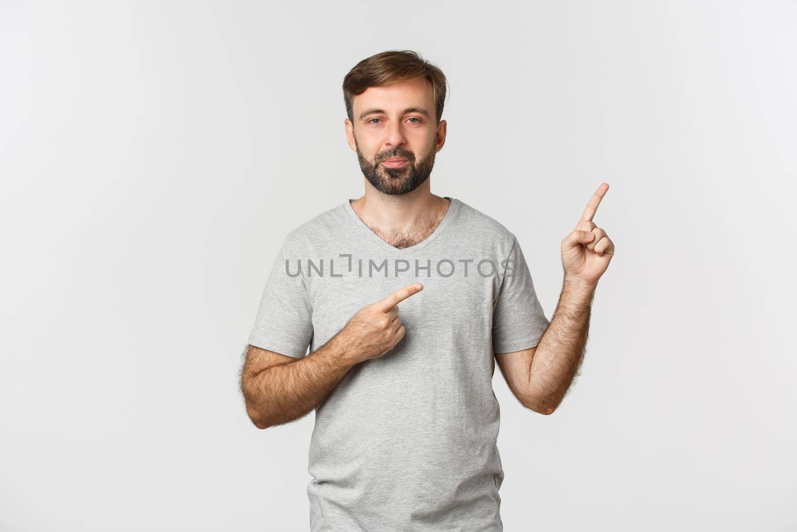Image of handsome bearded guy in gray t-shirt, showing logo, pointing fingers at upper right corner, standing over white background.