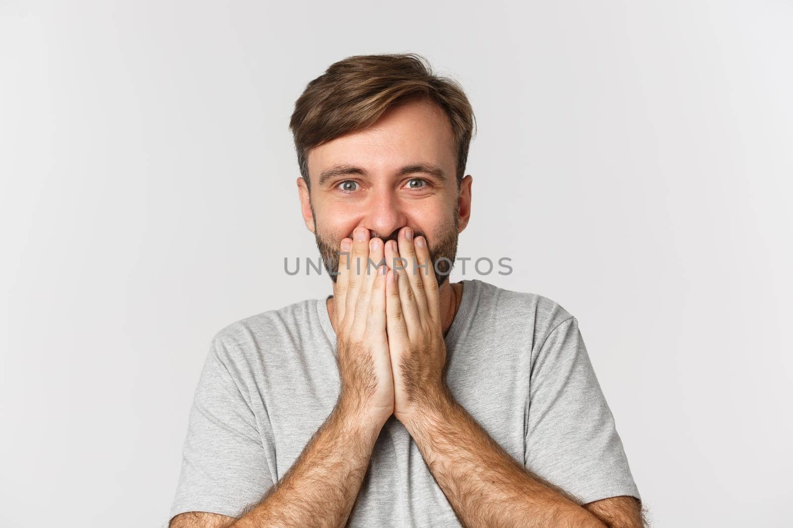 Close-up of excited and surprised man, cover mouth and giggle from amazement, standing over white background.