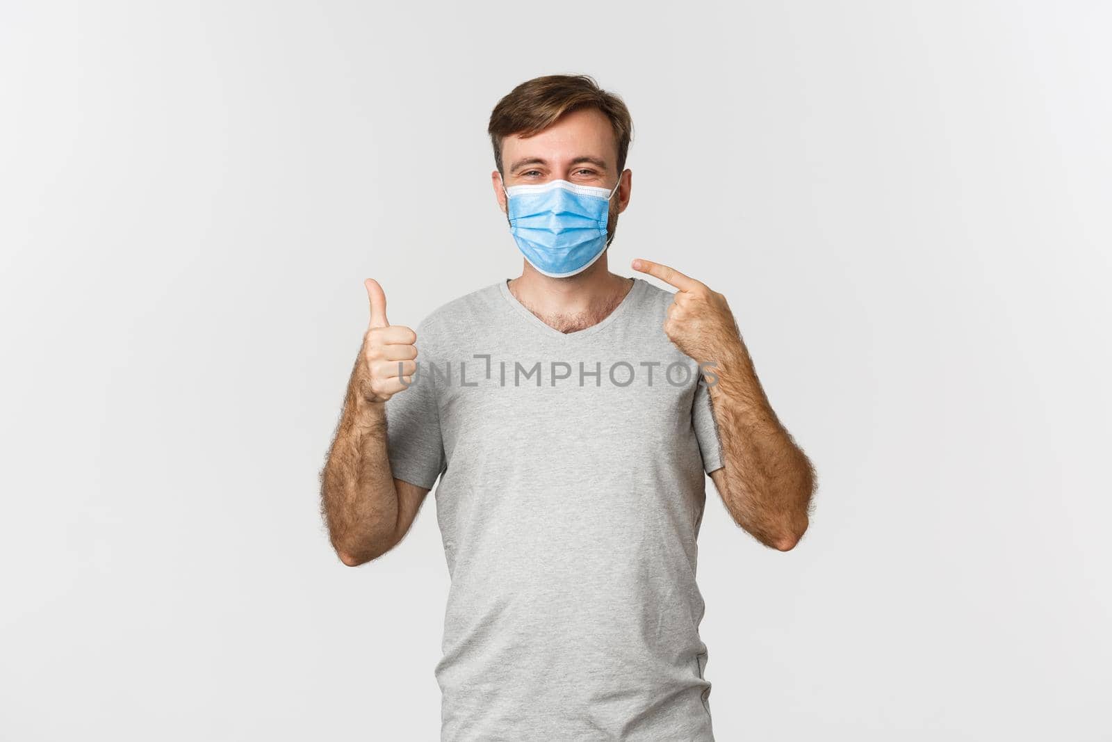 Concept of pandemic, covid-19 and social-distancing. Image of happy guy in gray t-shirt, pointing at medical mask and showing thumbs-up, standing over white background by Benzoix