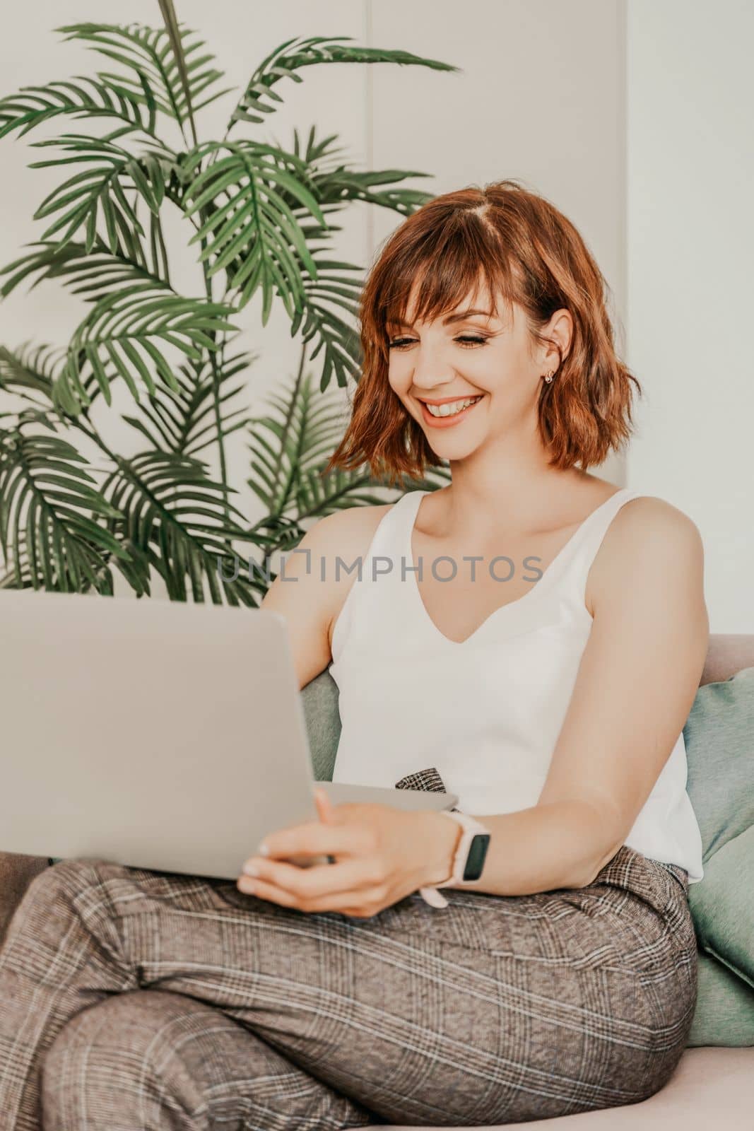 Brunette macbook sofa. Attractive young woman working on a laptop while sitting on the couch at home. She is wearing a light blouse and trousers