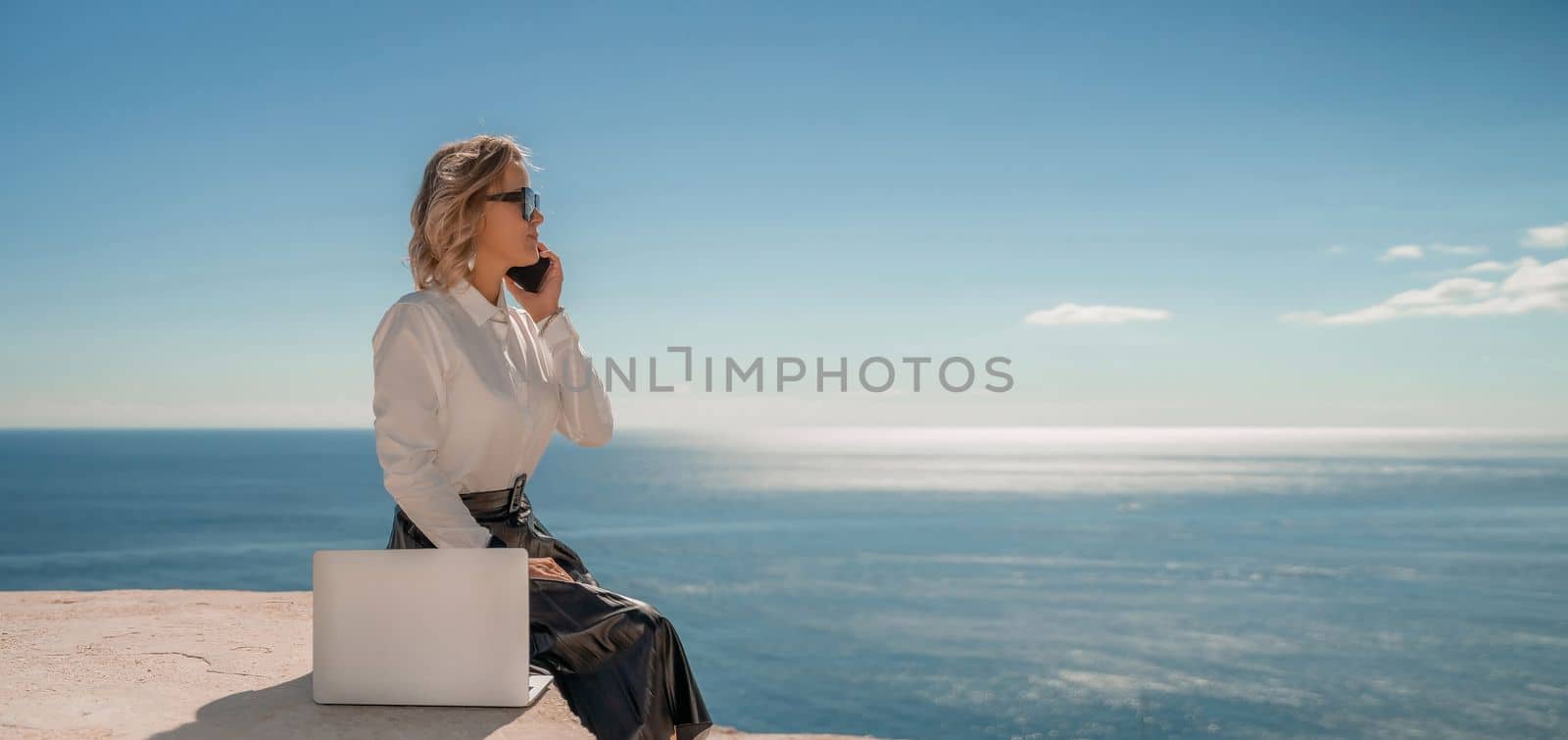 Business woman on nature in white shirt and black skirt. She works with an iPad in the open air with a beautiful view of the sea. The concept of remote work. by Matiunina