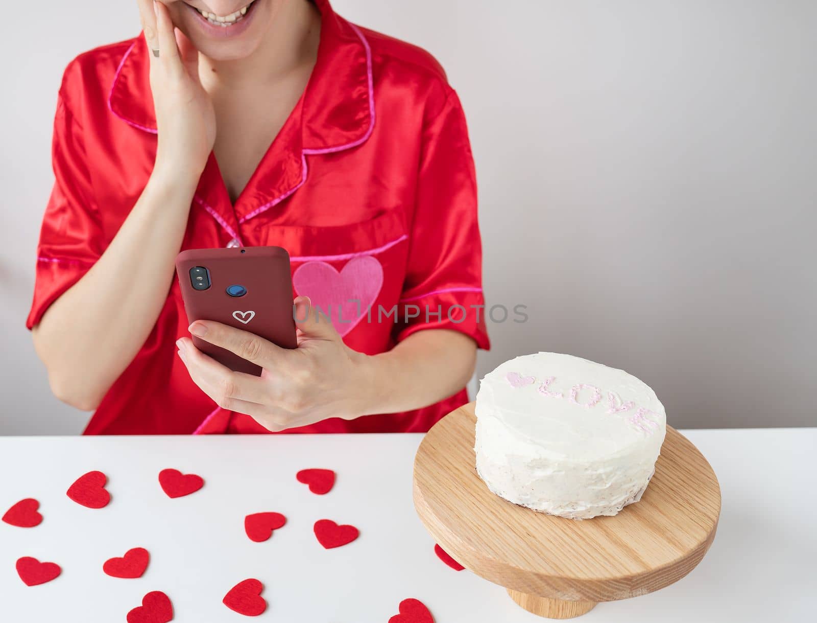 A happy girl in red clothes holds a smartphone in a cherry case with a heart and is happy with an SMS message from a loved one, a cake and small red hearts are on the table. Valentine's day and romantic date concept