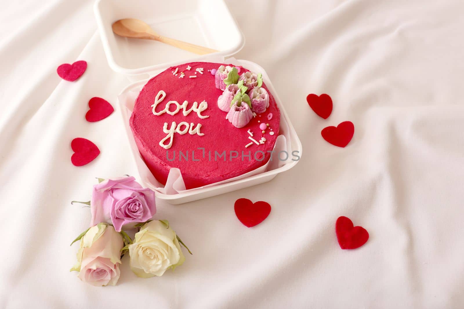 A plastic lunch box with a delicious red bento cake with the inscription Love you, with red hearts and roses lying on a white bed next to it. Close up. Copy space