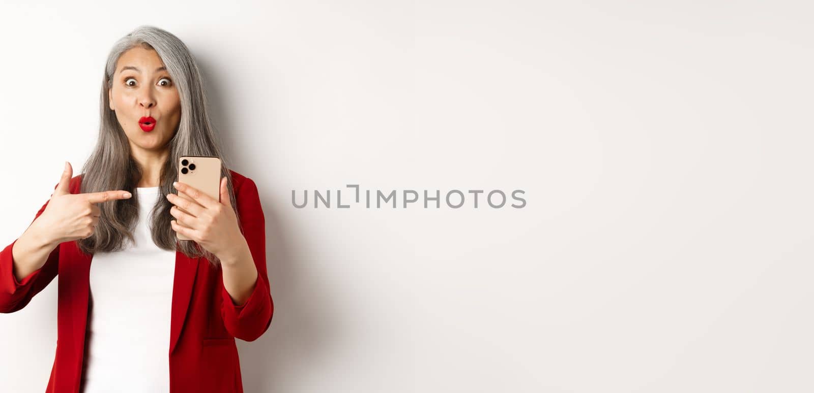 Amazed korean lady in red blazer pointing finger at mobile phone, looking curious at camera, white background.