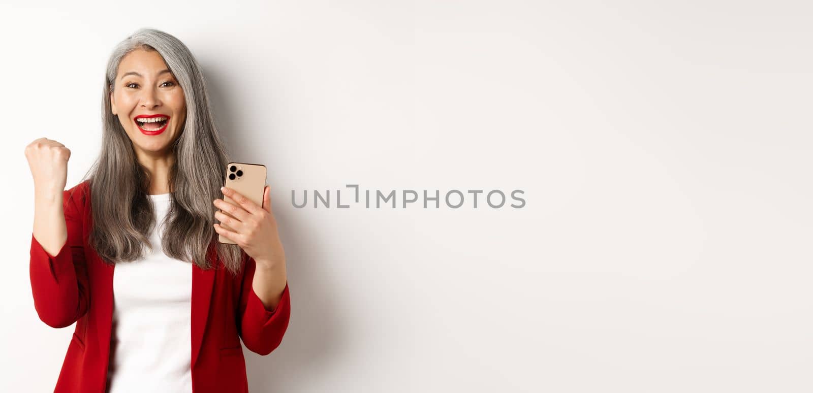 Asian old woman winning online, holding smartphone and making fist pump gesture to celebrate win, triumphing and smiling, standing over white background by Benzoix