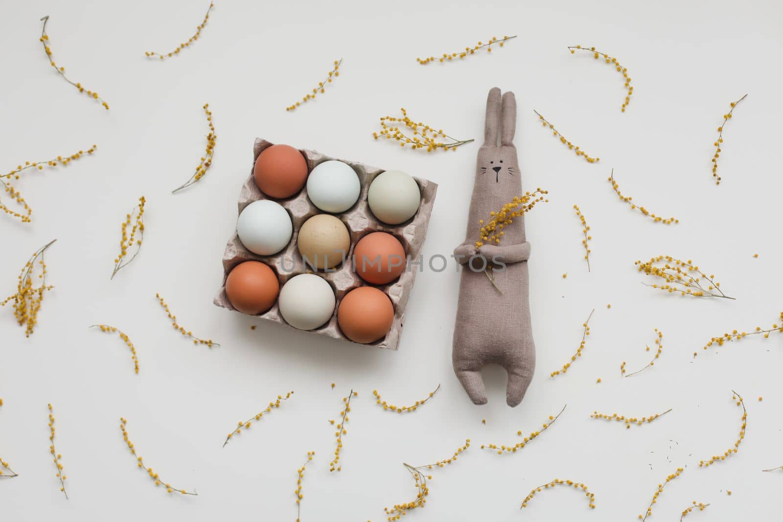 Funny handmade bunny rabbit toy with colorful chicken eggs and yellow mimosa flowers. Easter gift concept.