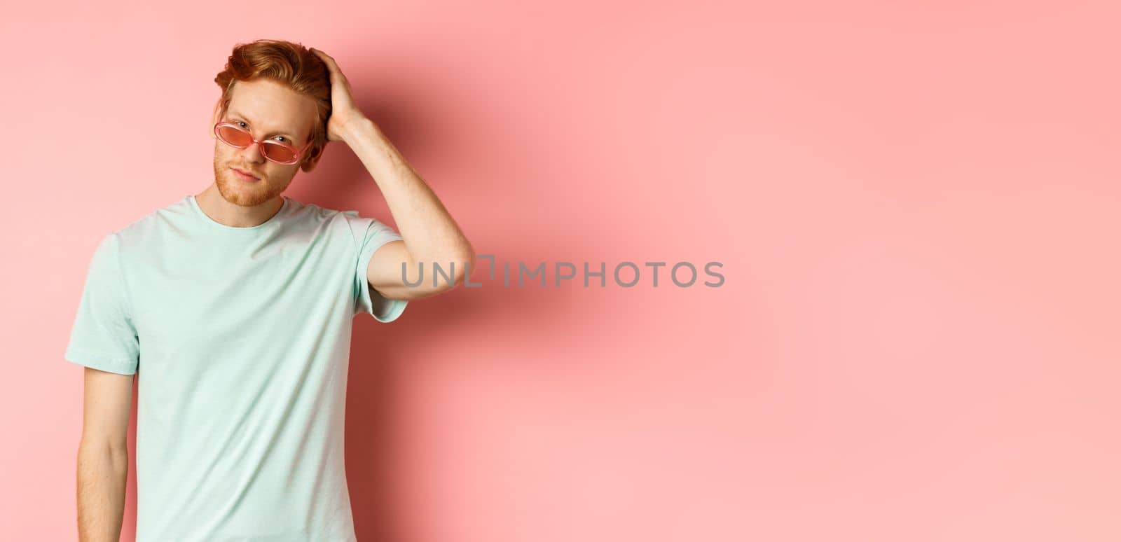 Handsome young redhead man in sunglasses, brushing hair with hand and looking smug and confident at camera, standing over pink background by Benzoix