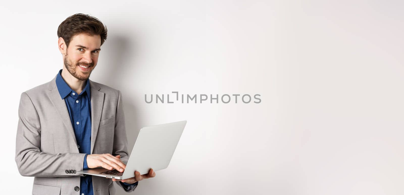 Successful smiling businessman working on laptop and looking happy at camera, standing in grey suit on white background by Benzoix