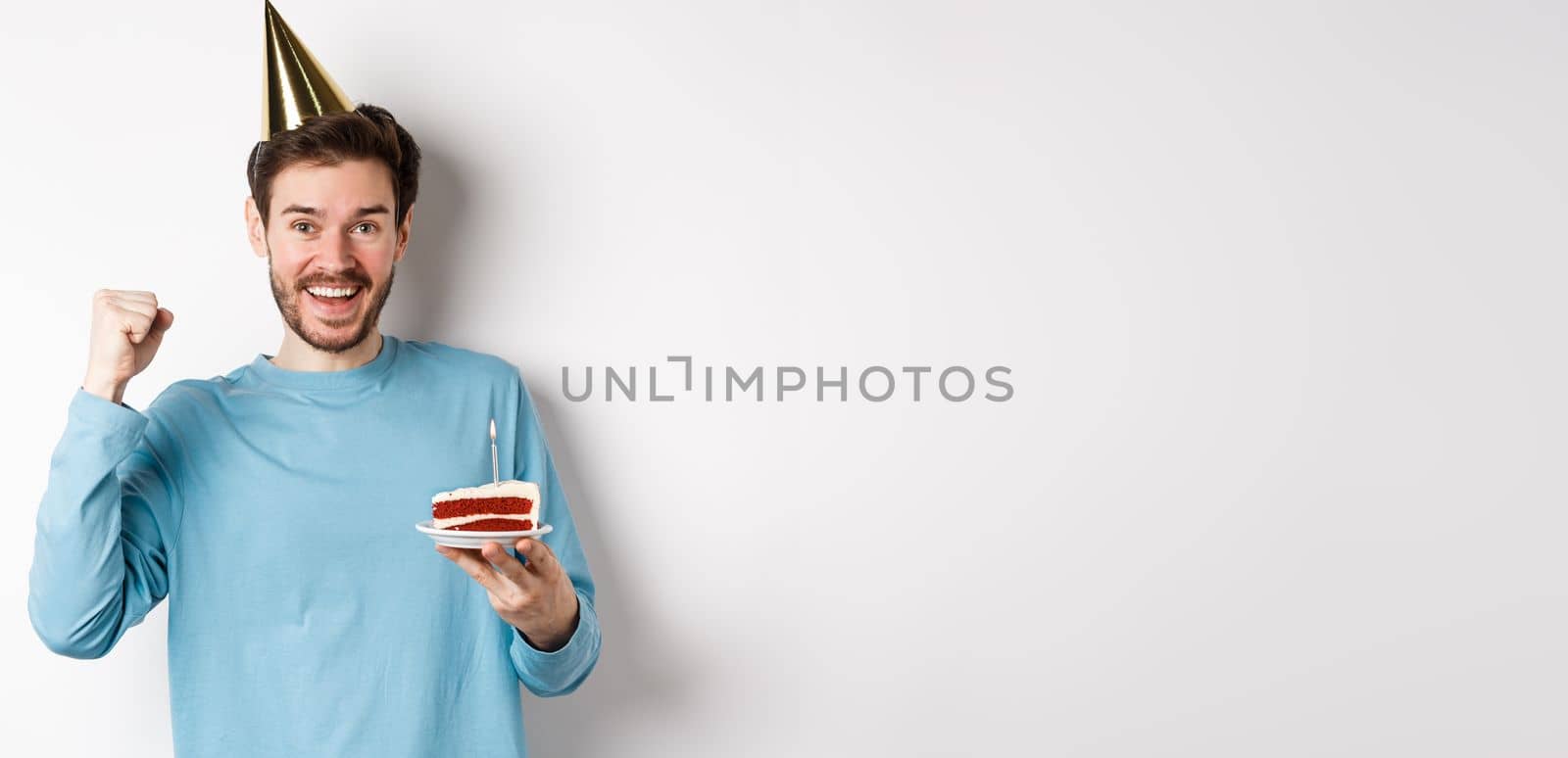 Celebration and holidays concept. Cheerful young man celebrating birthday in party hat, holding bday cake and looking happy, standing on white background.