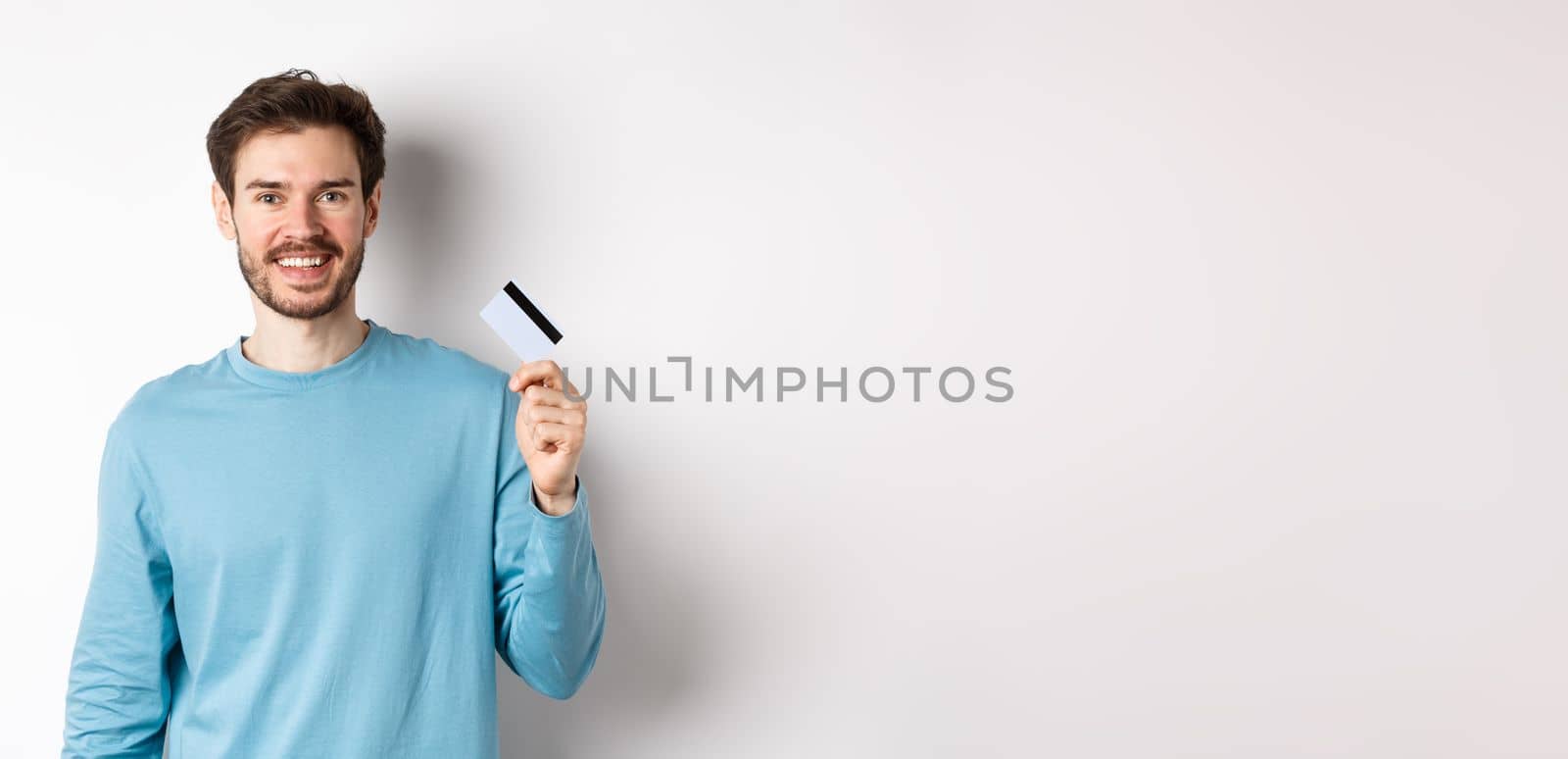Smiling male bank client showing plastic credit card with happy face, standing over white background.