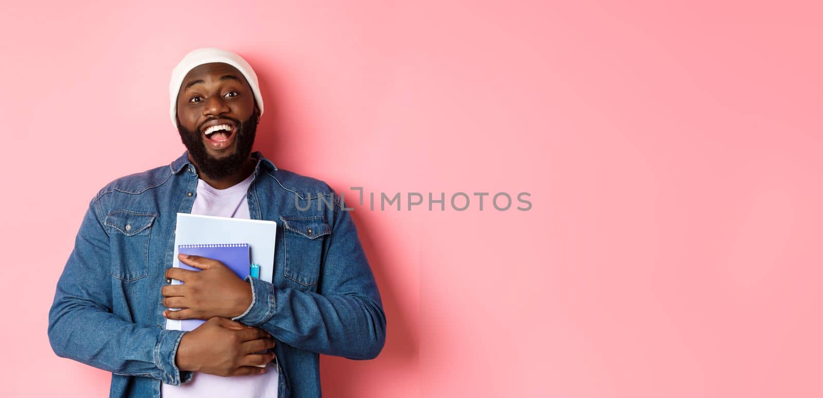 Education. Happy african-american male student in beanie holding notebooks, studying courses, smiling at camera, standing over pink background by Benzoix