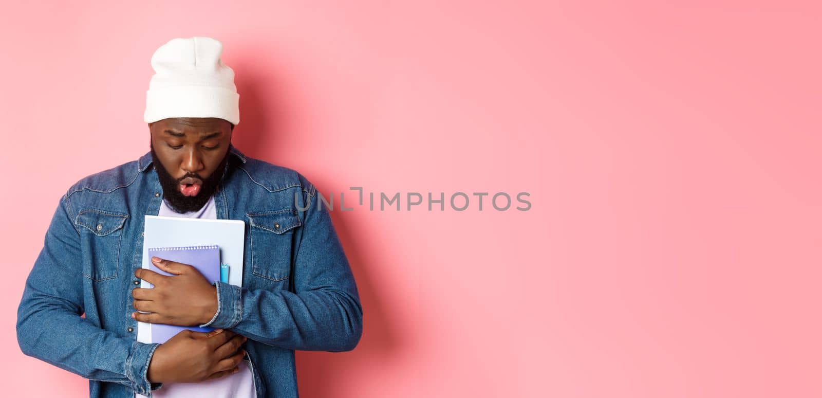 Education. Image of african-american bearded male student holding notebooks and looking down, drop something on floor, standing over pink background.