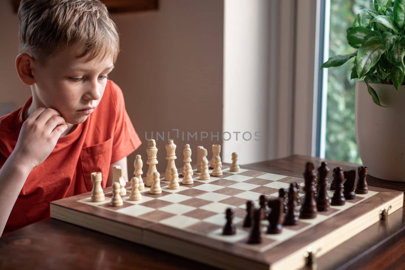 Young white child playing a game of chess on large chess board. Chess board on table in front of school boy thinking of next move by Len44ik