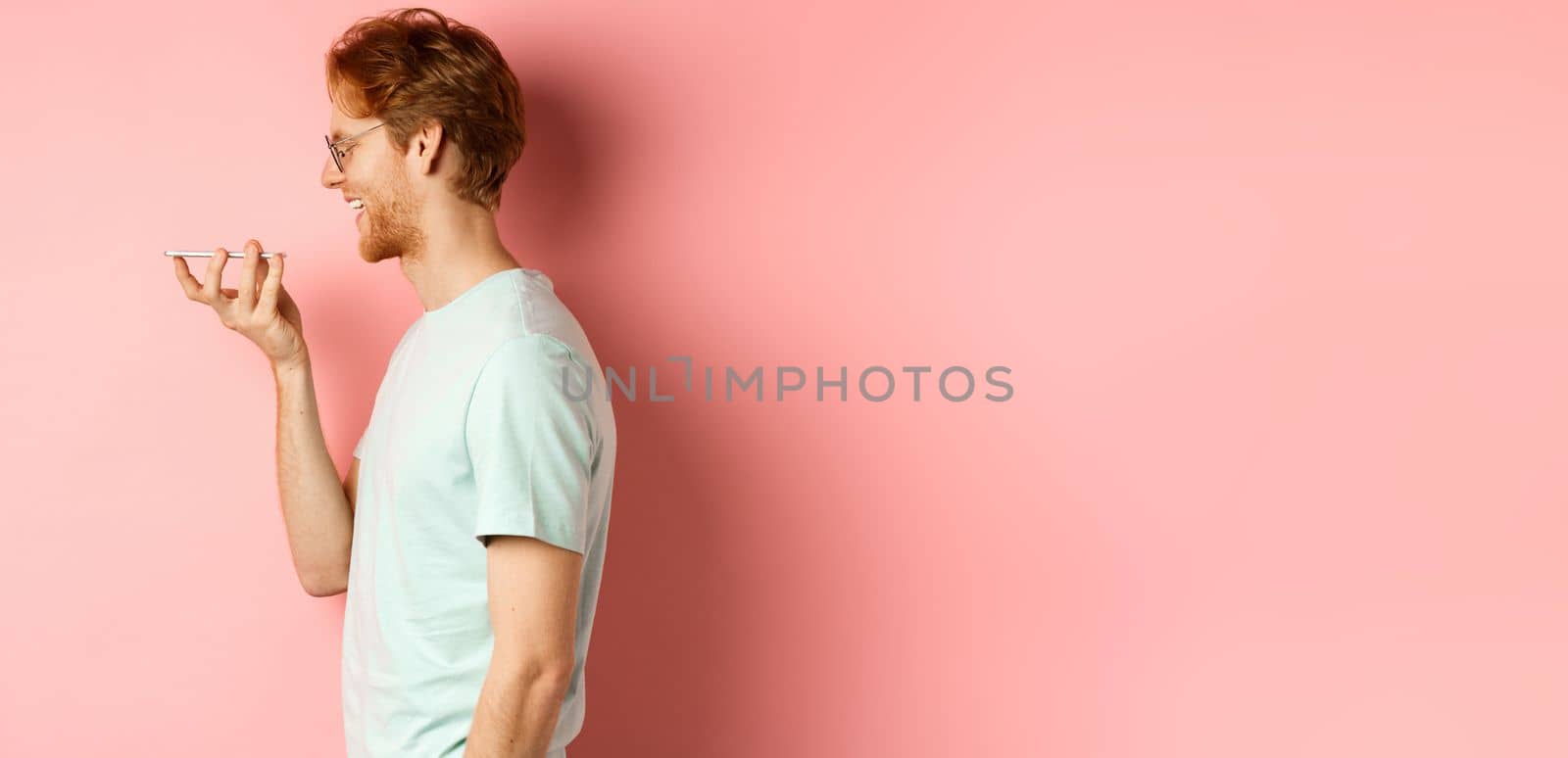 Profile portrait young man with red hair, smiling pleased while record voice message on smartphone, talking to virtual assistant, standing over pink background by Benzoix