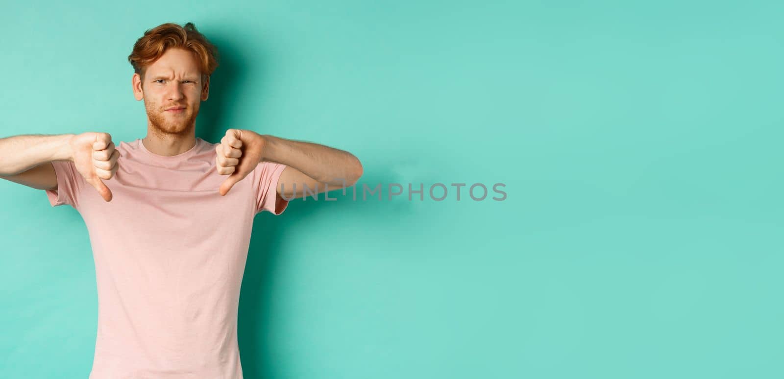 Disappointed guy with red hair showing thumbs-down, frowning and looking skeptical, epxress dislike and disapproval, standing over turquoise background.