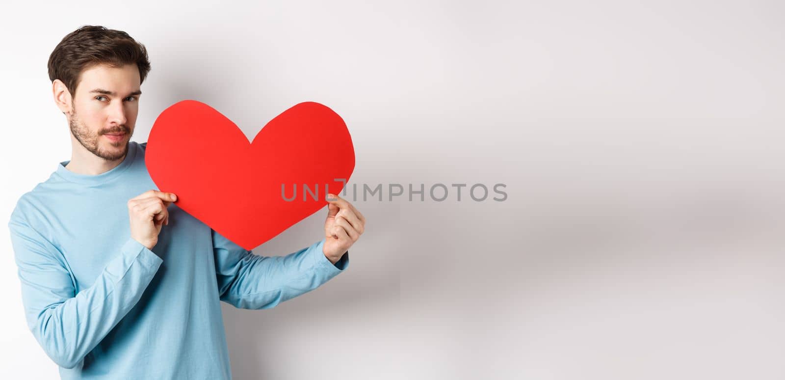 Valentines day. Handsome and romantic man holding big red valentine heart cutout, looking seductive at camera, making love confession, white background.