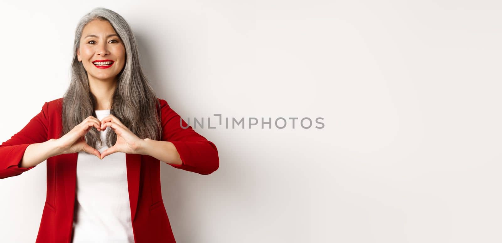 Beautiful asian mature woman in red blazer and makeup, showing heart sign and smiling, I love you gesture, standing over white background.