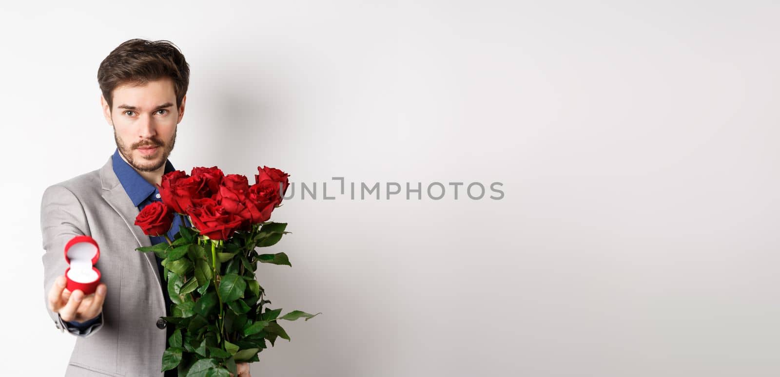 Handsome young man making a marriage proposal, stretch out hand with engagement ring and holding red roses, asking to marry him, looking confident at lover, white background by Benzoix