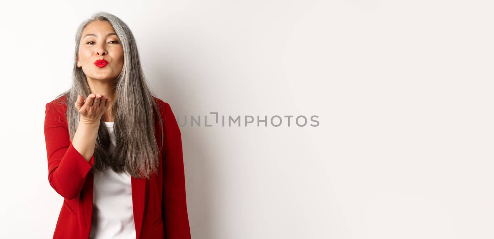 Elegant asian woman with red blazer and lips, blowing air kiss at camera, concept of valentines day and romance, white background.