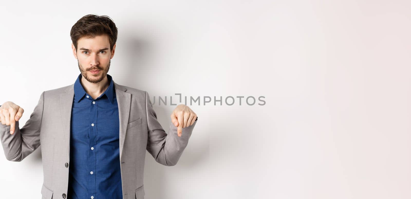 Look here. Handsome bearded man in suit pointing fingers down, looking confident at camera, showing banner, standing on white background.