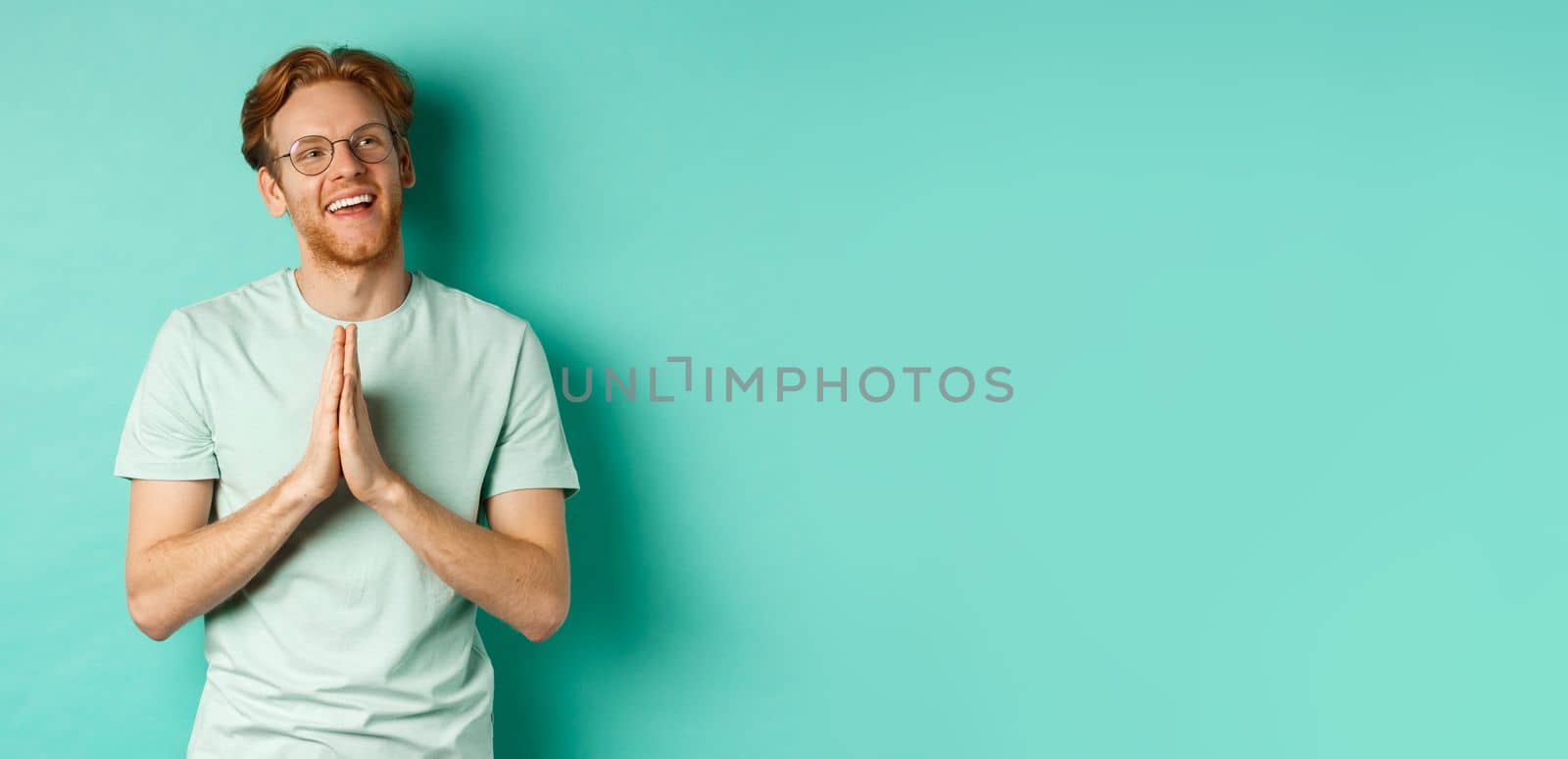 Hopeful redhead man with beard, wearing glasses and t-shirt, holding hands in namaste or plead gesture and looking right, smiling and thanking, standing over turquoise background by Benzoix