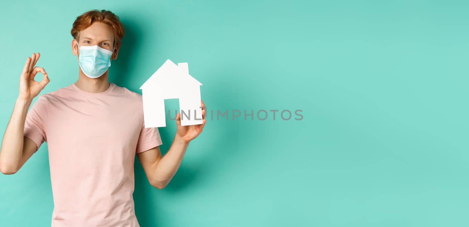Covid-19 and real estate concept. Cheerful guy in face mask showing house cutout and okay sign, standing over turquoise background.