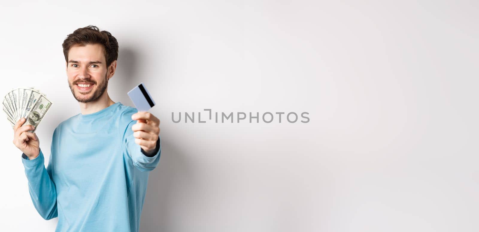 Smiling caucasian man holding money and giving you plastic credit card, standing on white background by Benzoix
