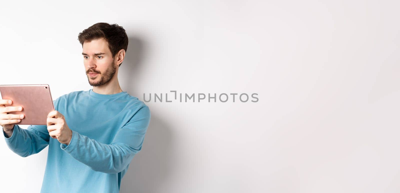Young man taking photo on digital tablet, looking serious at gadget screen, standing over white background by Benzoix