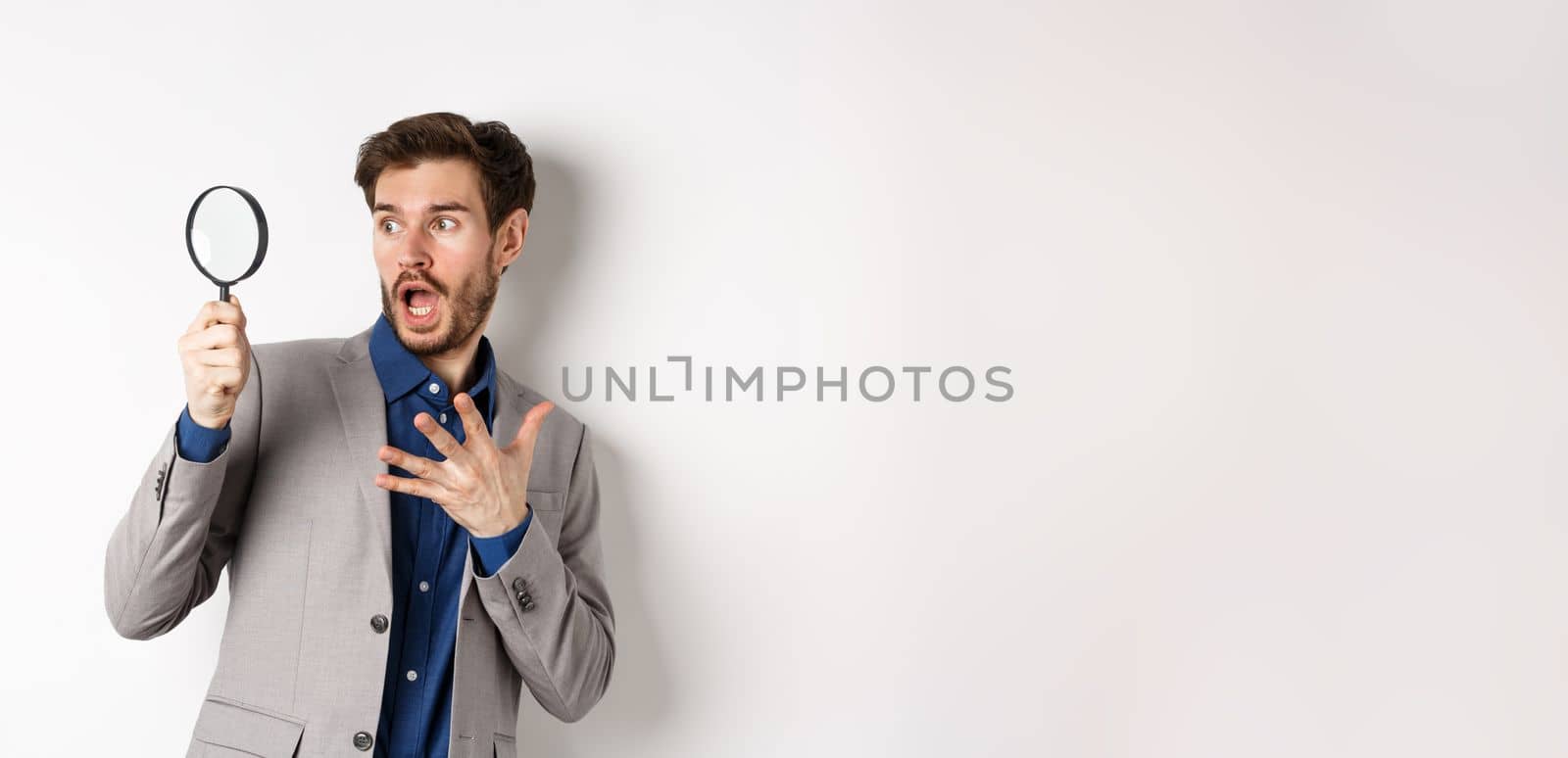 Shocked guy look through mafnifying glass with startled face, gasping and holding hand on chest scared, standing on white background.