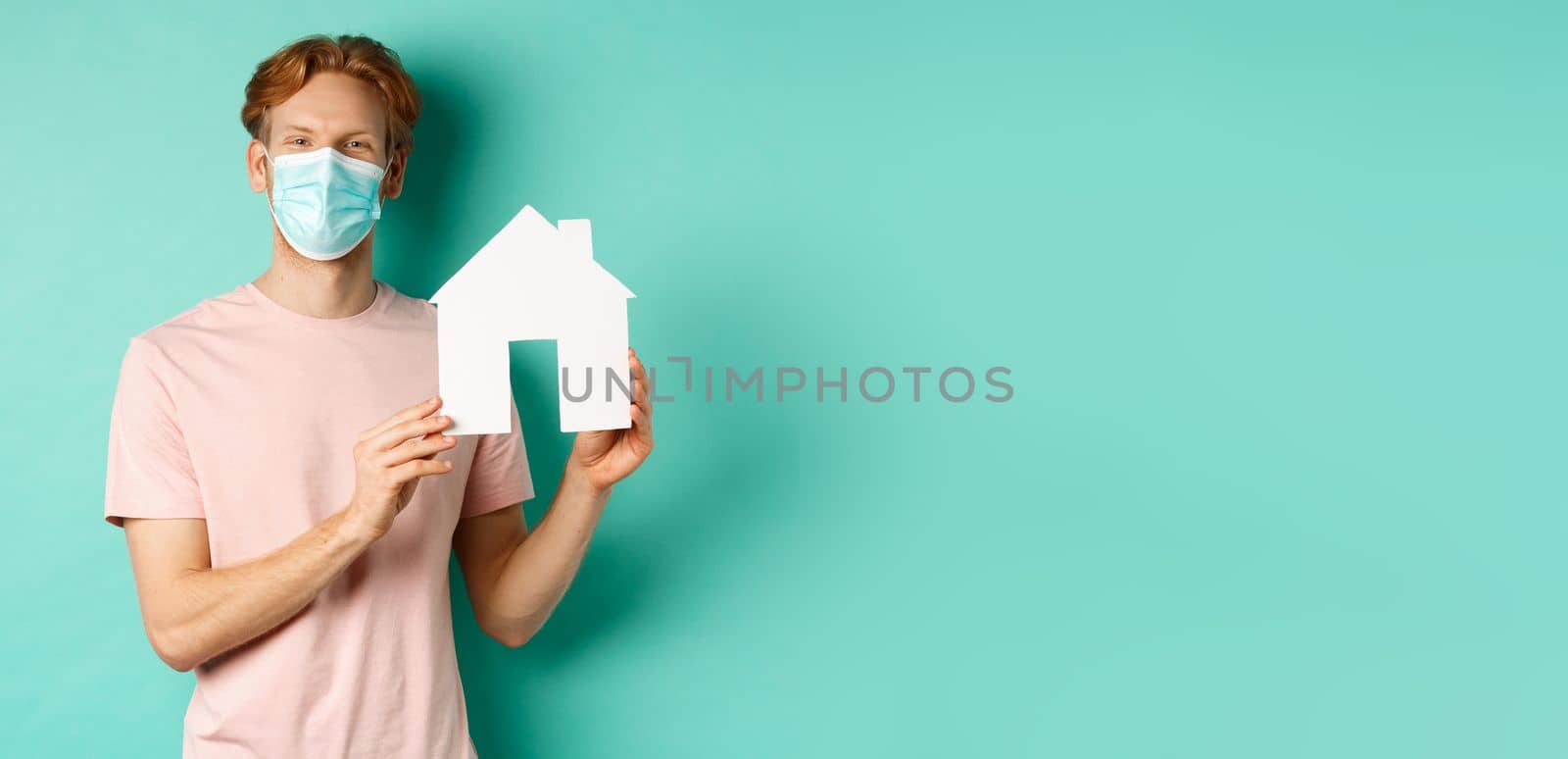 Covid-19 and real estate concept. Young happy man in face mask showing paper house cutout and smiling, offer property for sale, standing over mint background by Benzoix